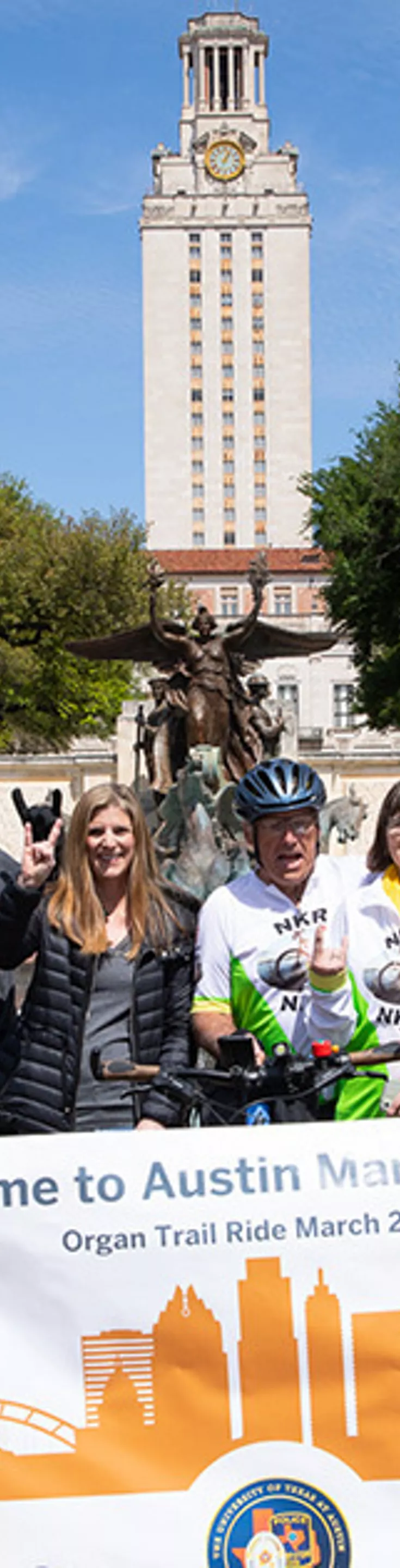 A diverse group of people, including cyclists and police officers on bikes, pose together outdoors in front of a university tower. The group holds a large banner that reads 'Welcome to Austin Mark and Lynn, Organ Trail Ride March 2023.' 