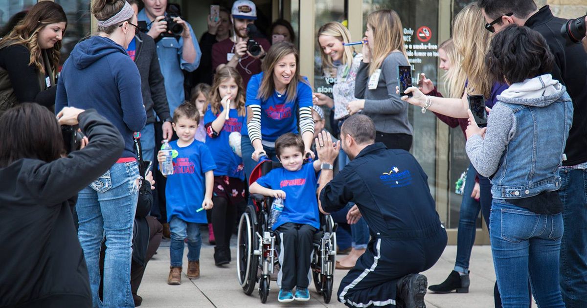 A group of children, some walking and one in a wheelchair, are greeted by a crowd outside a building. The children and several adults are wearing matching blue shirts, suggesting participation in a group event or cause. The scene is lively, with people taking photos and cheering, creating a supportive and celebratory atmosphere. The setting appears to be outdoors near a public entrance, with a diverse group of adults and children present.