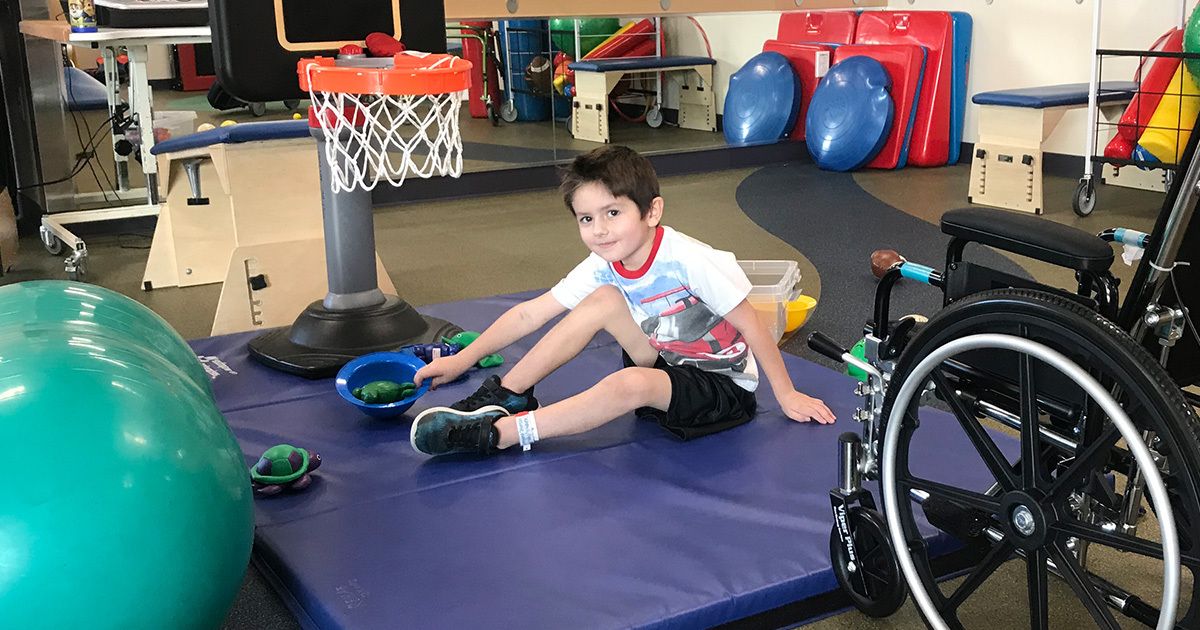 A young boy sits on a padded mat in a physical therapy or rehabilitation gym, reaching for rings near a small basketball hoop. The setting includes therapy equipment such as a large exercise ball, balance pads, and a wheelchair. The environment is bright and organized, designed for pediatric therapy activities. The boy is casually dressed and appears engaged in a therapeutic exercise.