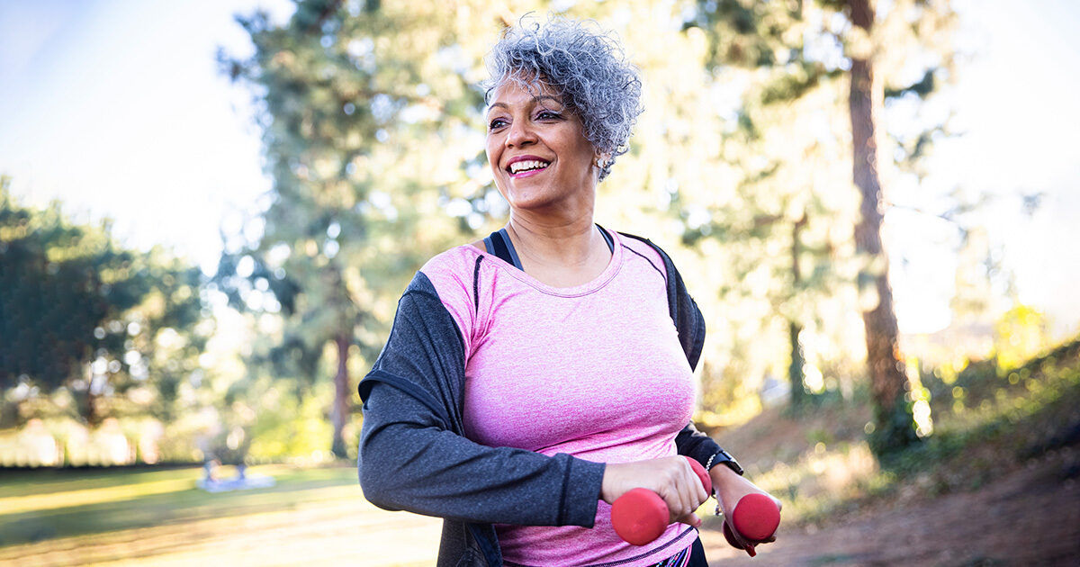 An older woman with short curly gray hair is seen exercising in a park setting, holding small red dumbbells. She wears a pink athletic top and a dark jacket, suggesting a casual fitness routine. The background features trees and sunlight, creating a bright and energetic atmosphere. The image conveys health, vitality, and an active lifestyle.