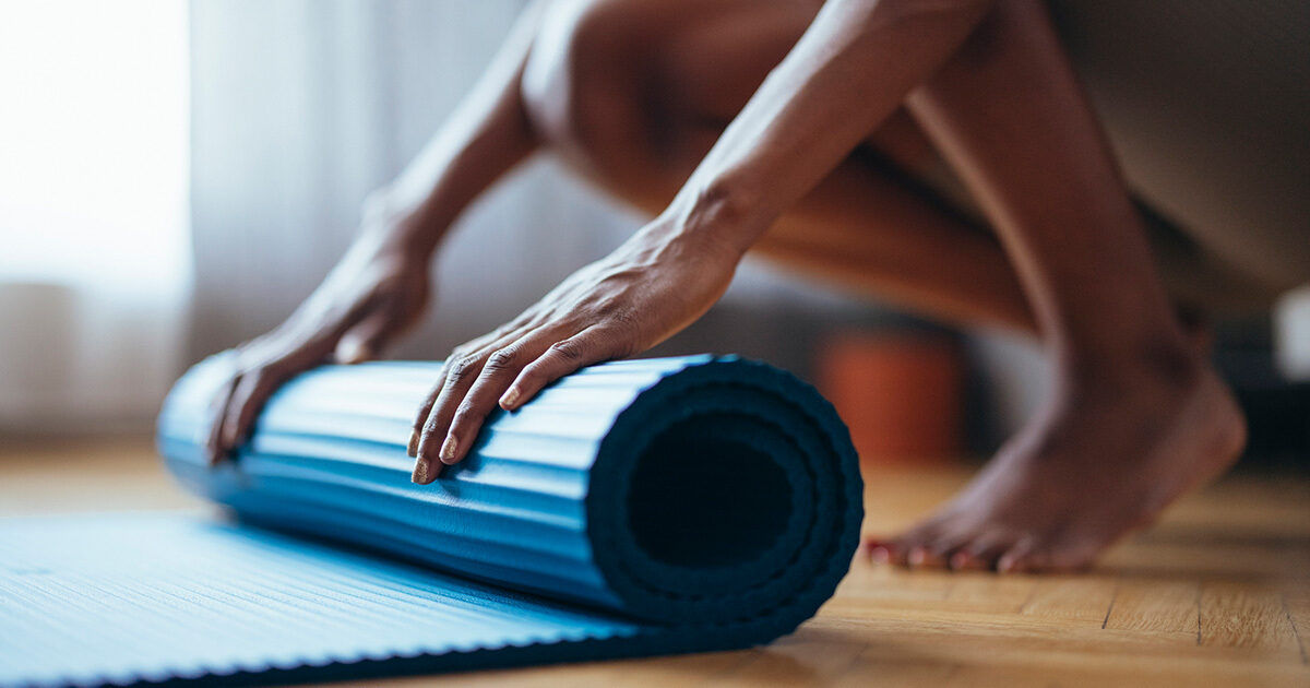 A close-up view shows a personâ€™s hands unrolling a blue yoga mat on a wooden floor. The scene suggests preparation for a fitness or yoga session in a home or studio setting. The image focuses on the mat and hands, with soft natural lighting and a calm, neutral palette. No visible text or numbers are present.