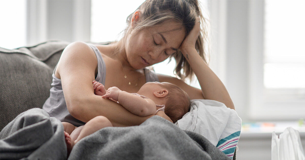 A woman is seated on a couch, breastfeeding a baby in a softly lit living room. She appears to be wearing casual, comfortable clothing and is partially covered by a blanket. The setting is calm and intimate, with natural daylight coming through the windows in the background. No visible text or numbers are present in the image.