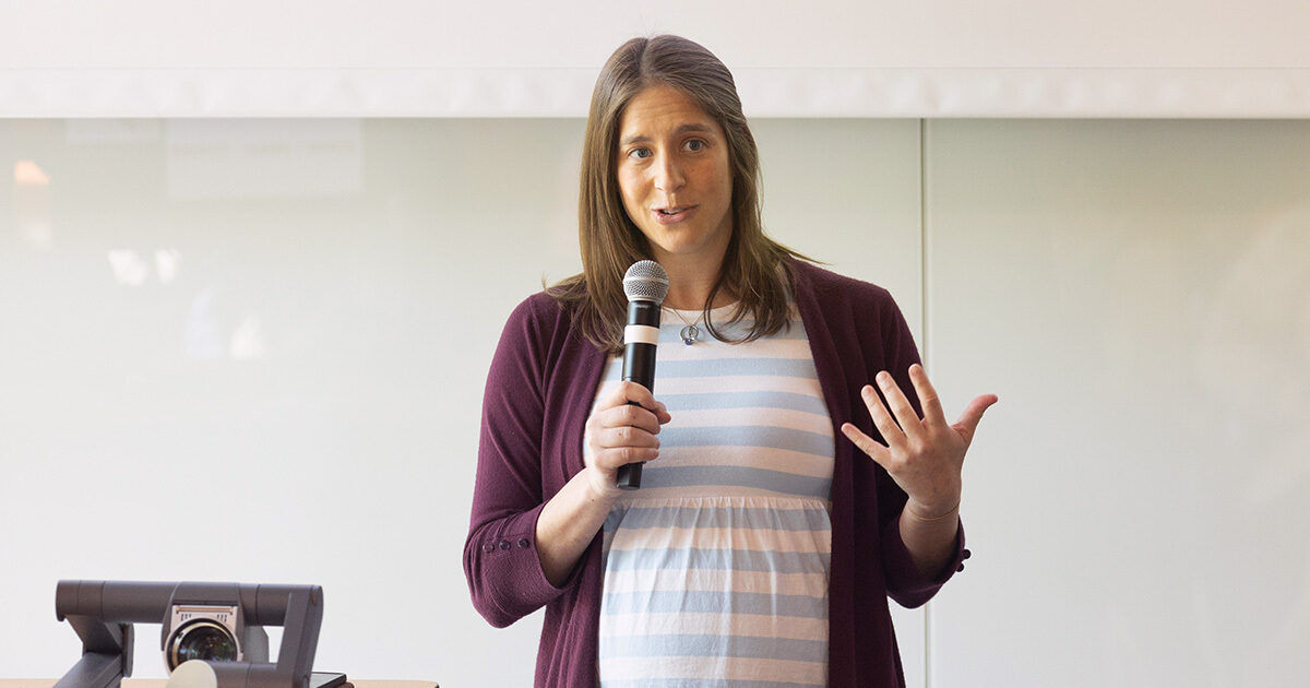 A woman stands in front of a light-colored wall, holding a microphone and gesturing with her hand. She is wearing a striped dress and a maroon cardigan. A projector is visible on a table to her left, suggesting a presentation or lecture setting. The atmosphere is professional and focused, with soft natural lighting.
