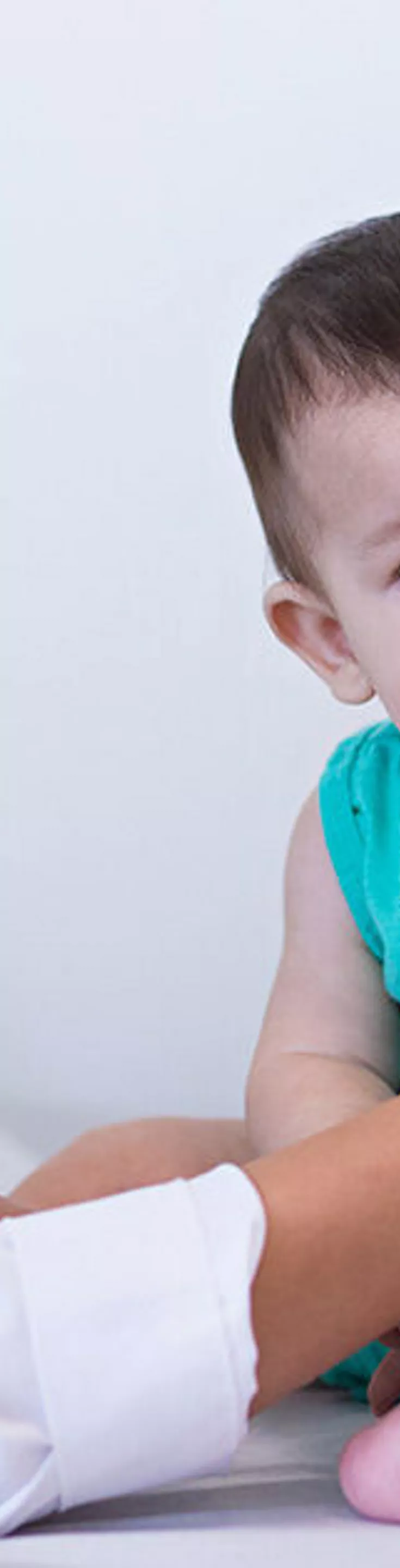A healthcare professional uses a stethoscope to examine a baby sitting on an examination table. The setting is a bright, clean medical office with framed pictures on the wall. The baby is wearing a turquoise outfit and appears calm during the checkup. The scene conveys a sense of care and professionalism in a pediatric healthcare environment.