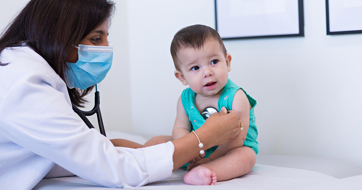 A healthcare professional uses a stethoscope to examine a baby sitting on an examination table. The setting is a bright, clean medical office with framed pictures on the wall. The baby is wearing a turquoise outfit and appears calm during the checkup. The scene conveys a sense of care and professionalism in a pediatric healthcare environment.