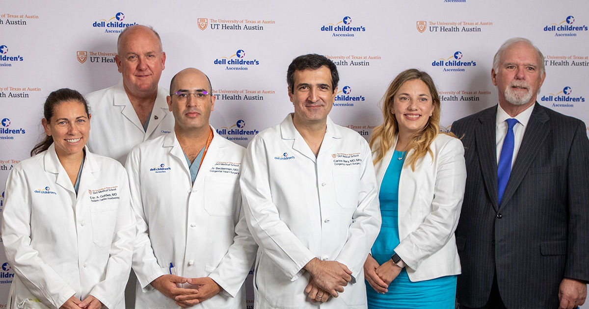 A group of medical professionals stands together in front of a branded backdrop featuring 'dell children's' and 'UT Health Austin' text. Several individuals are wearing white lab coats, while one person is in a suit and another in a teal dress with a white blazer. The setting appears formal, likely at a conference or hospital event, with a focus on healthcare and teamwork.