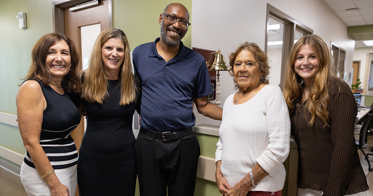A group of five adults stands together in a hallway, posing for a photo. The setting appears to be indoors, possibly in a healthcare or office environment, with a bell mounted on the wall in the background. The individuals are dressed in casual and business-casual attire, and the mood is friendly and supportive. The hallway features light green walls and doors leading to other rooms.