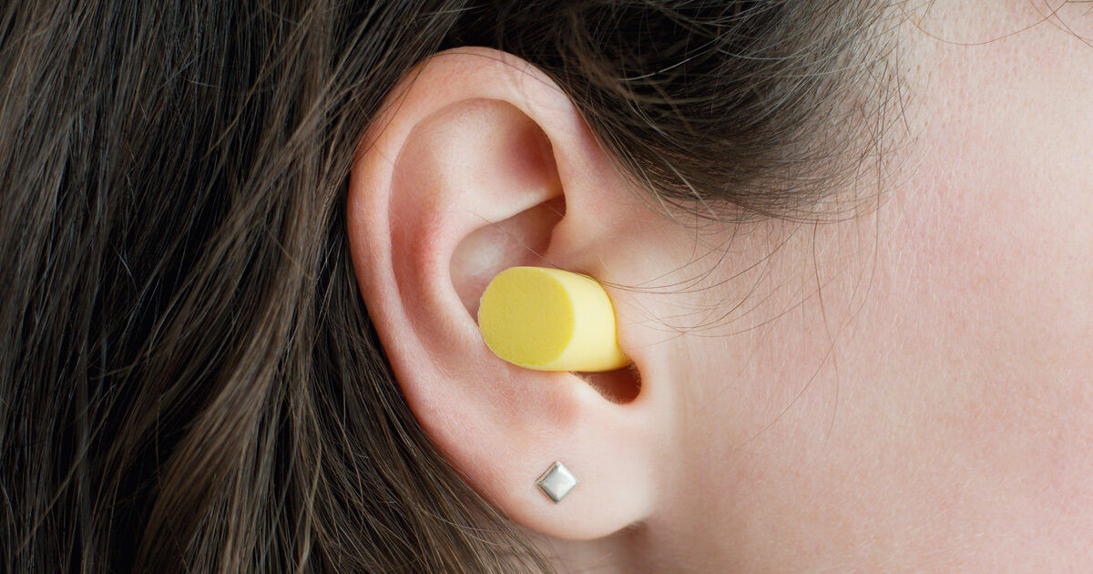 A yellow foam earplug is inserted into the ear of a woman with brown hair. The image is a close-up, focusing on the ear and the earplug, with a small silver square earring visible on the earlobe. The background is neutral, highlighting the earplug and earring details.