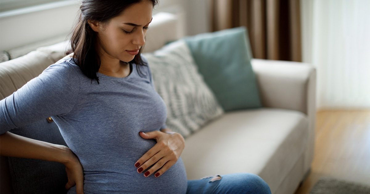 A pregnant woman sits comfortably on a beige sofa in a modern living room, gently cradling her belly with both hands. She is wearing a casual long-sleeve blue shirt and dark pants. The setting features soft natural light, neutral tones, and a calm, intimate atmosphere. The focus is on the expectant mother and the peaceful home environment.