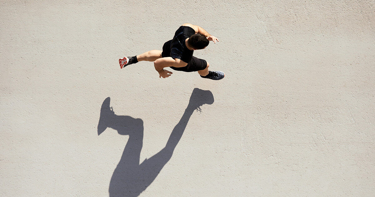 A single athlete is captured mid-stride from an overhead perspective, running on a light concrete surface. The runner's dynamic pose and extended limbs create a dramatic, elongated shadow on the ground. The scene is minimalistic, with a neutral palette and a focus on movement and energy. The athlete appears to be an adult male, dressed in black athletic wear.