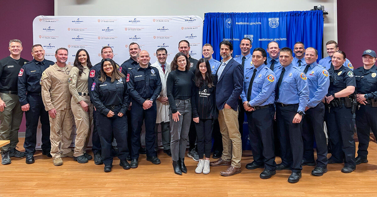 A large group of emergency responders, including police officers, firefighters, and officials, pose together indoors. The setting features branded step-and-repeat and blue curtain backdrops, suggesting a formal event or ceremony. The group includes both men and women of various ages, all in uniform or business attire. The mood is positive and professional, with everyone smiling for the camera. Some visible patches and insignias are present on uniforms, but no specific brands or numbers are clearly identifiable.