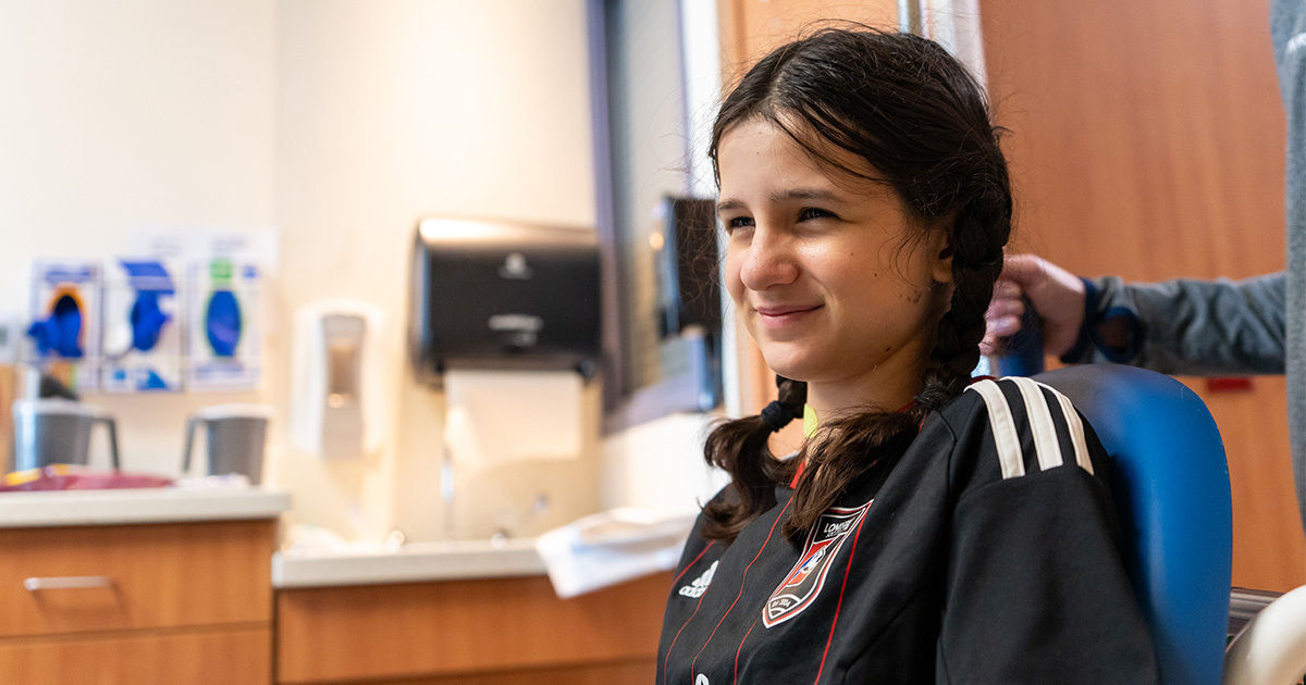 A young individual with braided hair sits in a blue chair inside a medical or dental clinic. The person is wearing a black sports jacket with white stripes and a visible crest on the chest. The setting includes a countertop, paper towel dispenser, and medical posters on the wall, suggesting a healthcare environment.