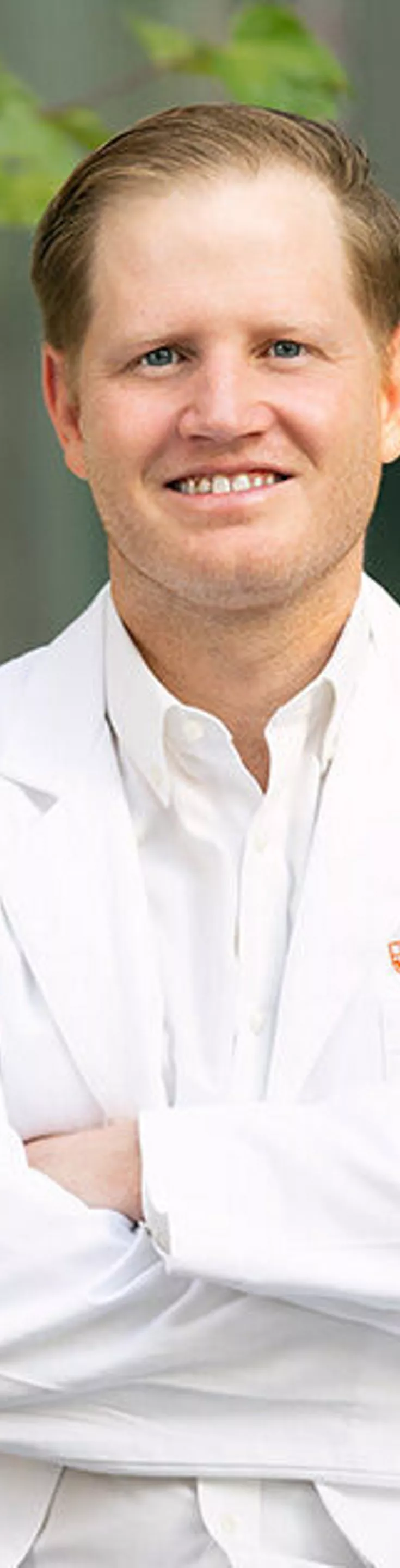 A person wearing a white medical coat stands with arms crossed outside, next to a modern building with glass windows. The setting includes green foliage and a neutral-toned wall, suggesting a healthcare or academic environment. The coat features visible embroidered text on the chest, though specific details are not fully legible. The overall mood is professional and composed.