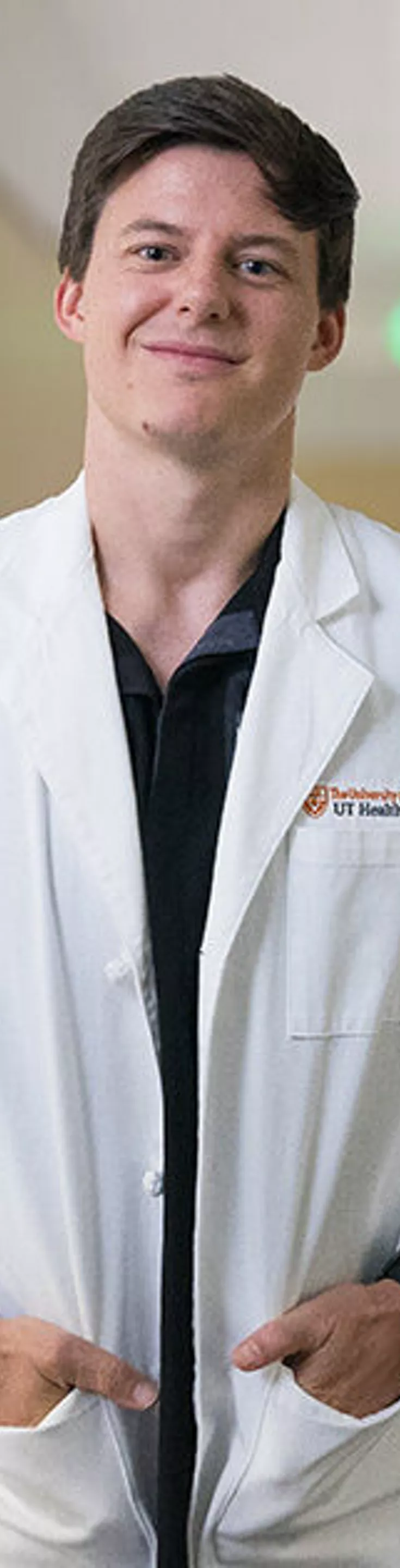 A person in a white lab coat stands confidently in a well-lit optometry clinic. The setting features advanced eye examination equipment and a clean, organized workspace with neutral tones. The individual appears to be a professional, possibly an optometrist, ready for patient care. The environment suggests a focus on vision health and clinical precision.