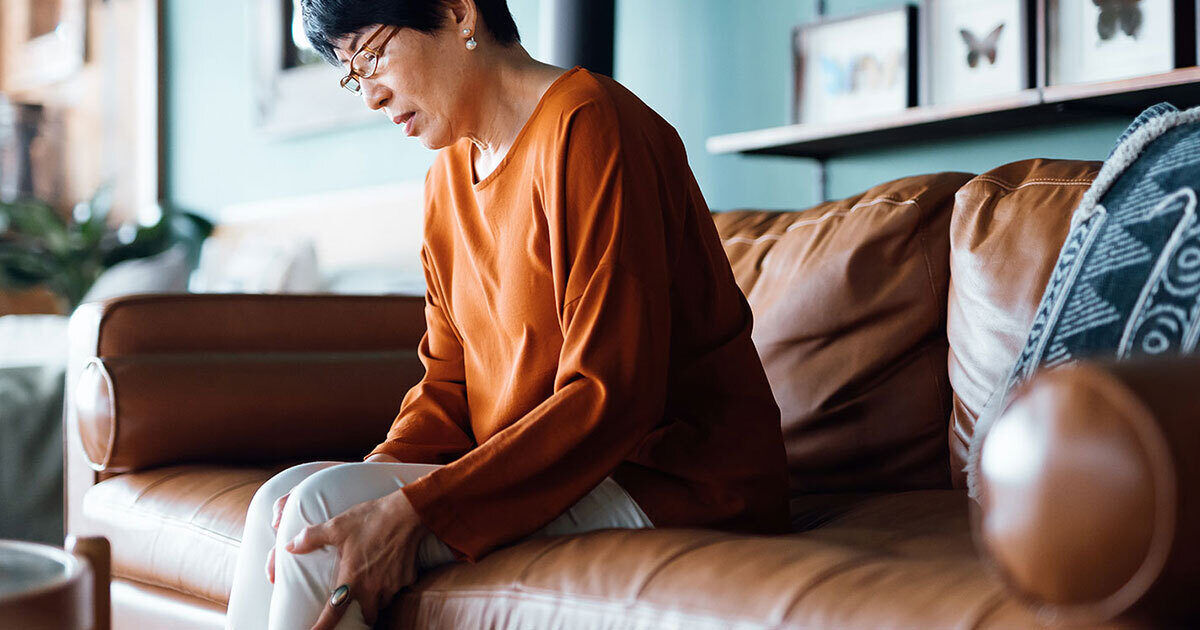 An adult sits on a brown leather sofa in a modern living room. The individual is wearing a burnt orange long-sleeve top and light-colored pants. The setting features decorative pillows, framed wall art, and a soft, natural light ambiance. The overall mood is calm and relaxed, with a contemporary interior style.