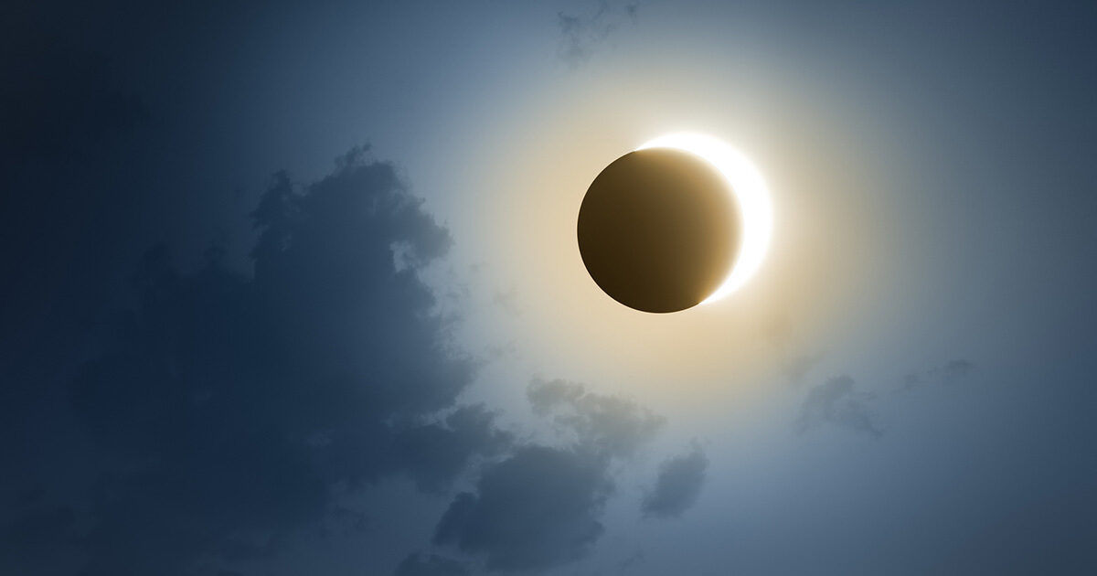A striking solar eclipse is captured as the moon passes directly in front of the sun, creating a glowing corona effect. Wispy clouds partially obscure the sky, adding depth and atmosphere to the celestial event. The scene is bathed in a cool, ethereal light, emphasizing the contrast between the darkened moon and the bright solar halo. No visible text or numbers are present in the image.