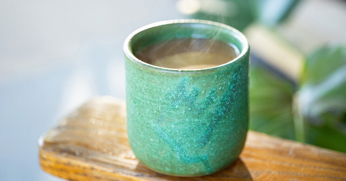 A green ceramic cup filled with a steaming hot beverage sits on a wooden surface. The setting appears to be outdoors, with soft natural light and blurred greenery in the background. The cup's textured glaze and earthy tones create a calm, rustic mood. No visible text or numbers are present in the image.