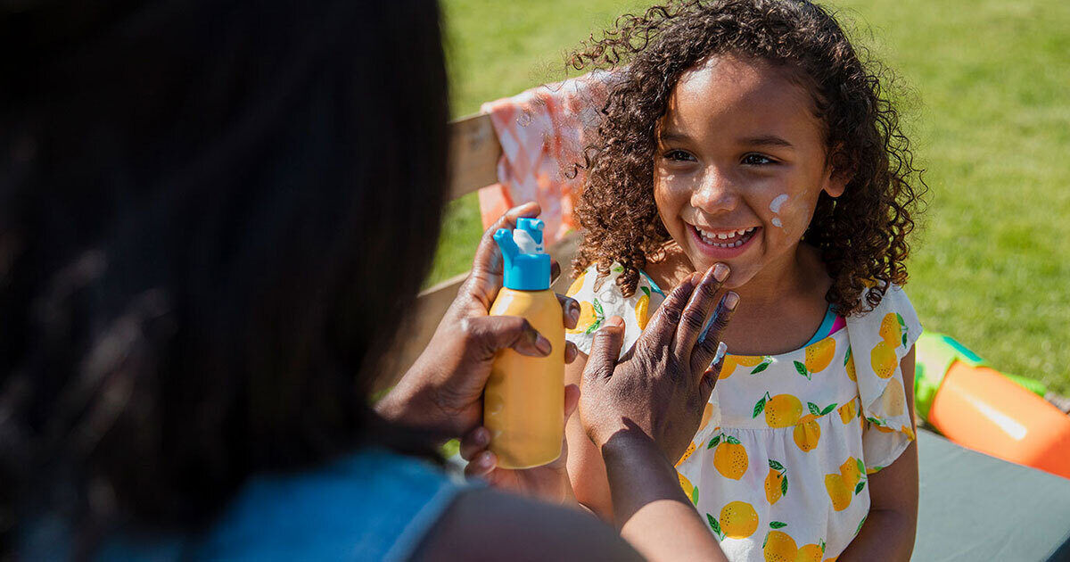 An adult is applying sunscreen lotion to a young child outdoors. The child is wearing a white dress with a colorful fruit pattern and is standing in a grassy area, possibly a park or backyard. The sunscreen bottle is yellow with a blue cap, and the scene is brightly lit, suggesting a sunny day. The focus is on sun protection and outdoor activity.