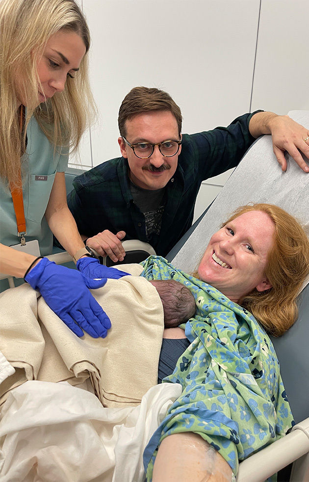 Katharina holds newborn Elliott in a hospital bed, smiling with her husband and a care team member beside her.