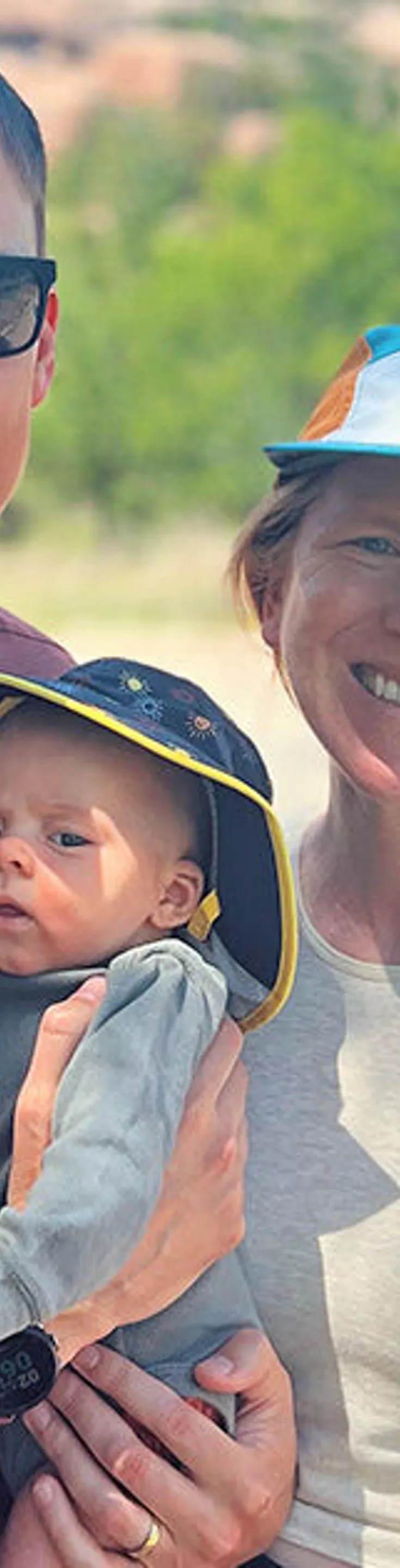 A group of three people, including two adults and a child, stand together outdoors on a sunny day. The adults are dressed in casual, sporty clothing, with one wearing a cap and the other a smartwatch. The background features greenery and a road, suggesting a park or nature setting. The overall mood is relaxed and cheerful, with bright, natural lighting.