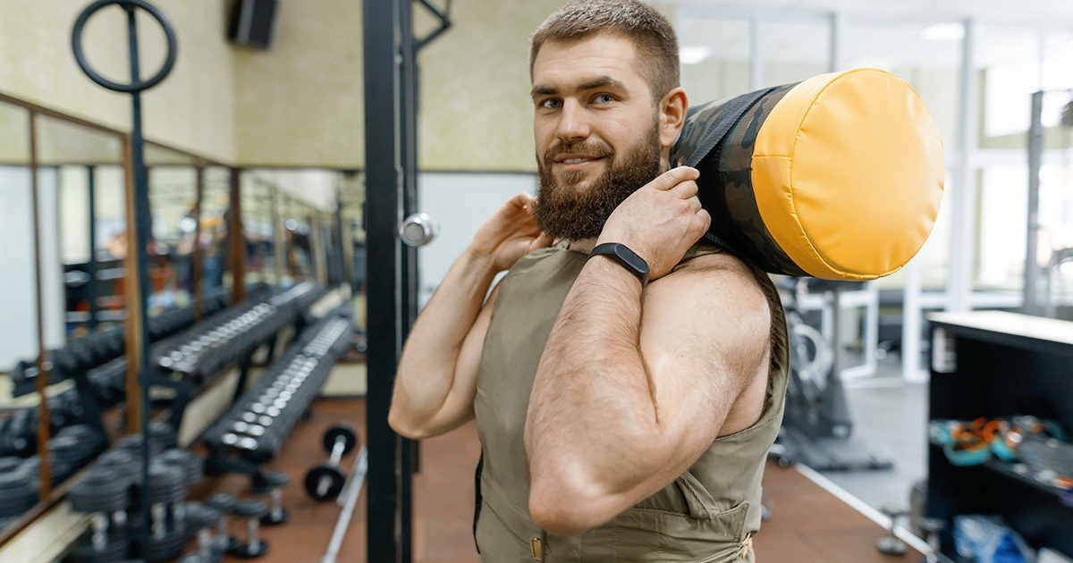 A man is performing a strength training exercise with a yellow and black weighted sandbag on his shoulders. The scene is set in a well-equipped gym with mirrors, dumbbells, and various fitness equipment visible in the background. He is wearing a sleeveless top and a fitness tracker on his wrist. The environment is brightly lit, emphasizing a focused and active mood.
