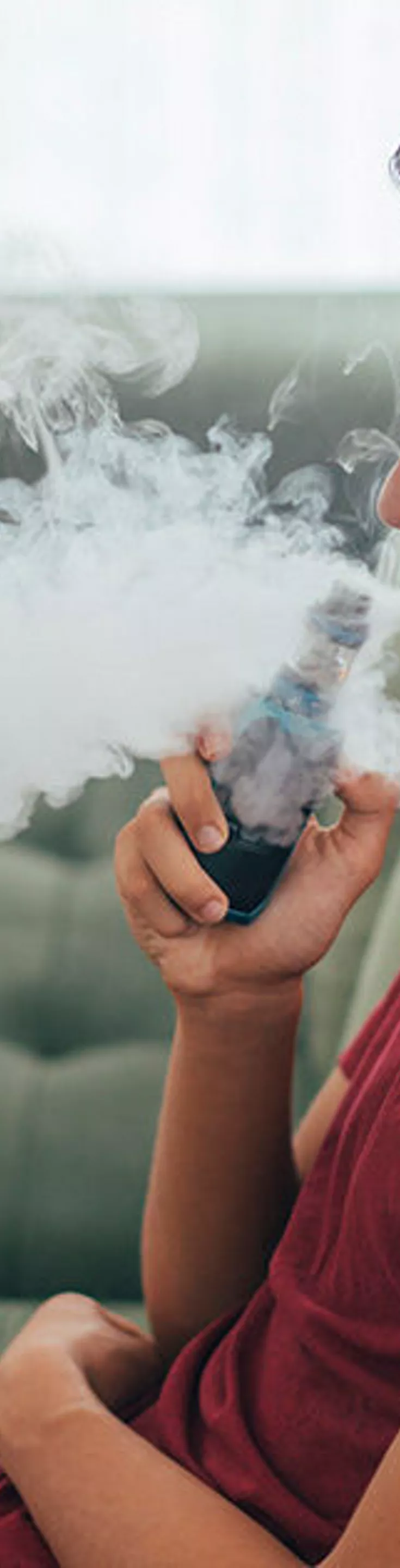 A young individual is seated on a green sofa, exhaling a large cloud of vapor from an electronic vaping device. The setting appears to be a modern living room with soft natural light and comfortable furniture. The person is wearing a red t-shirt and casual attire, contributing to a relaxed and informal atmosphere. No visible text or numbers are present in the image.