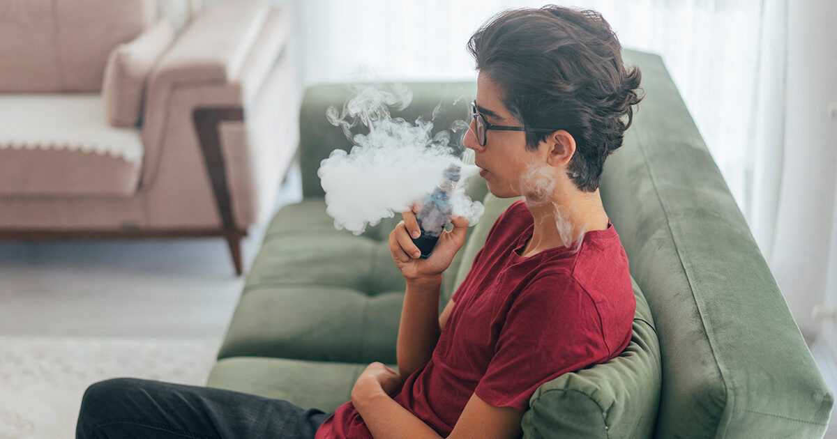 A young individual is seated on a green sofa, exhaling a large cloud of vapor from an electronic vaping device. The setting appears to be a modern living room with soft natural light and comfortable furniture. The person is wearing a red t-shirt and casual attire, contributing to a relaxed and informal atmosphere. No visible text or numbers are present in the image.