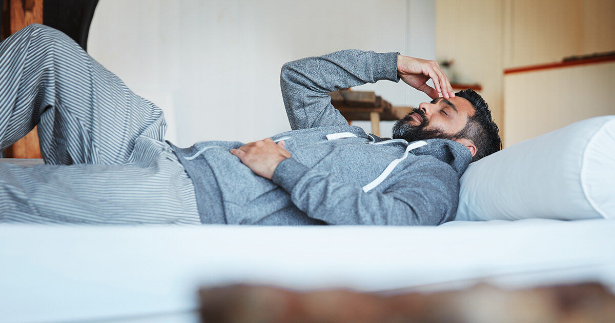 An adult male is lying on a bed, dressed in a grey hoodie and striped pajama pants. The setting appears to be a cozy bedroom with soft lighting and a relaxed atmosphere. The man is resting with one hand on his stomach and the other near his head, suggesting a moment of rest or contemplation. The overall mood is calm and casual, with neutral tones dominating the scene.