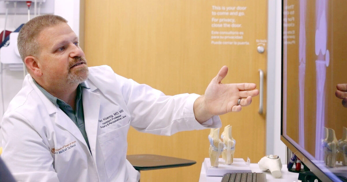 A medical professional in a white lab coat gestures toward a computer screen displaying a knee x-ray. The setting is a modern examination room with anatomical knee models visible on the desk. The environment is clinical and professional, with clear text instructions on the door in the background. The overall mood is informative and focused, with neutral tones dominating the palette.