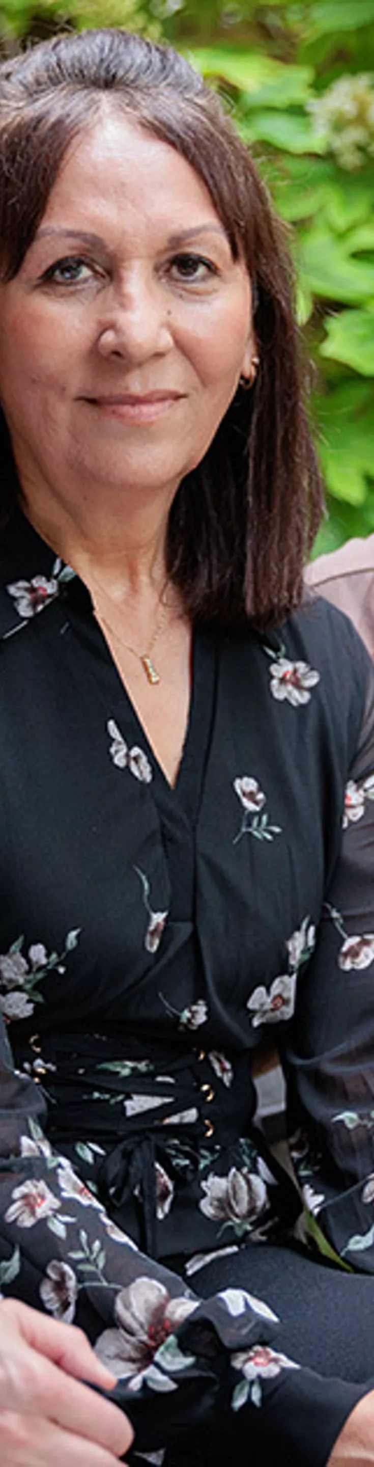 Three adult women are seated together on a black wooden bench in an outdoor courtyard. One wears a white coat, another a floral blouse, and the third a brown zip-up top with a circular emblem. The background features greenery and a building wall, creating a relaxed and professional atmosphere.