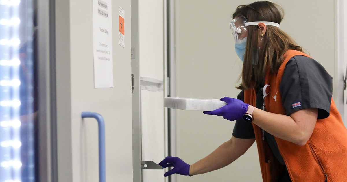 A person wearing an orange vest and purple gloves is placing a tray into a cold storage unit or freezer. The setting appears to be a laboratory or medical facility, with visible signage and equipment. The individual is engaged in a careful handling activity, suggesting the storage of sensitive materials. The environment is clean and professional, with neutral tones and a focus on safety.