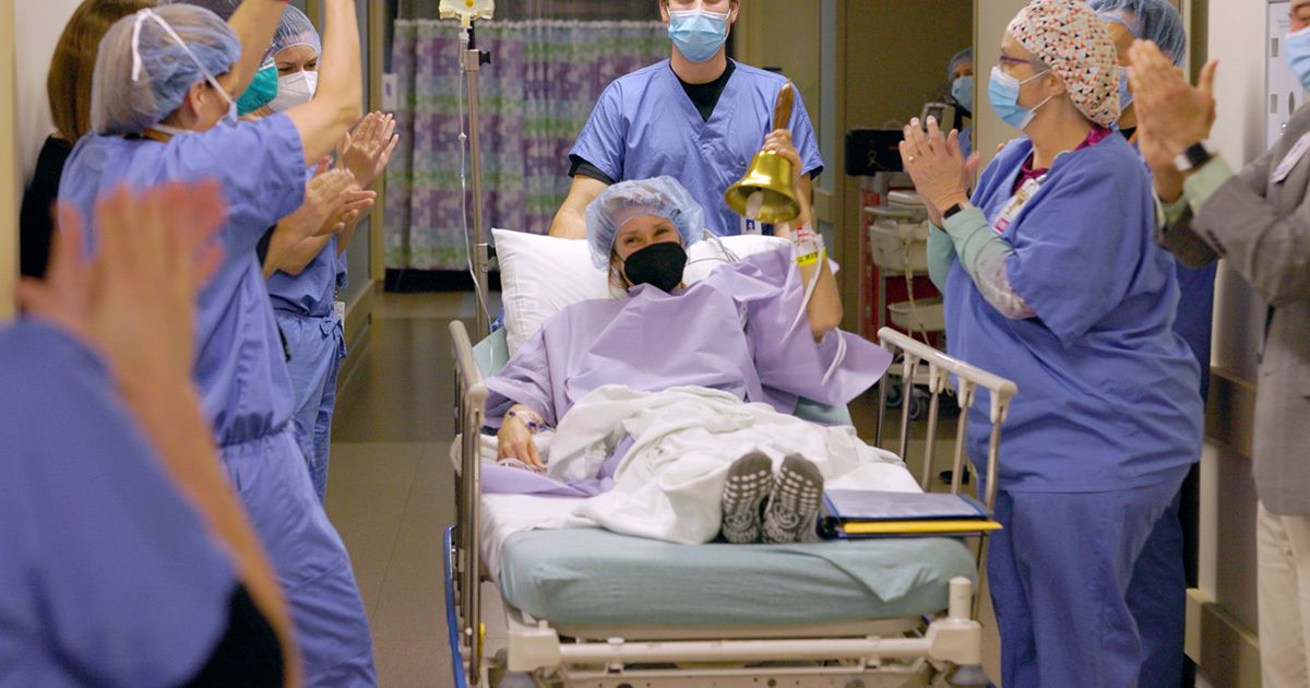 A patient in a hospital bed is being wheeled down a hallway, ringing a bell in celebration. The patient and surrounding medical staff are wearing masks and scrubs, indicating a healthcare setting. Staff members are clapping, creating a supportive and joyful atmosphere. The scene takes place indoors, with bright lighting and a sense of achievement. No visible text or numbers are present in the image.
