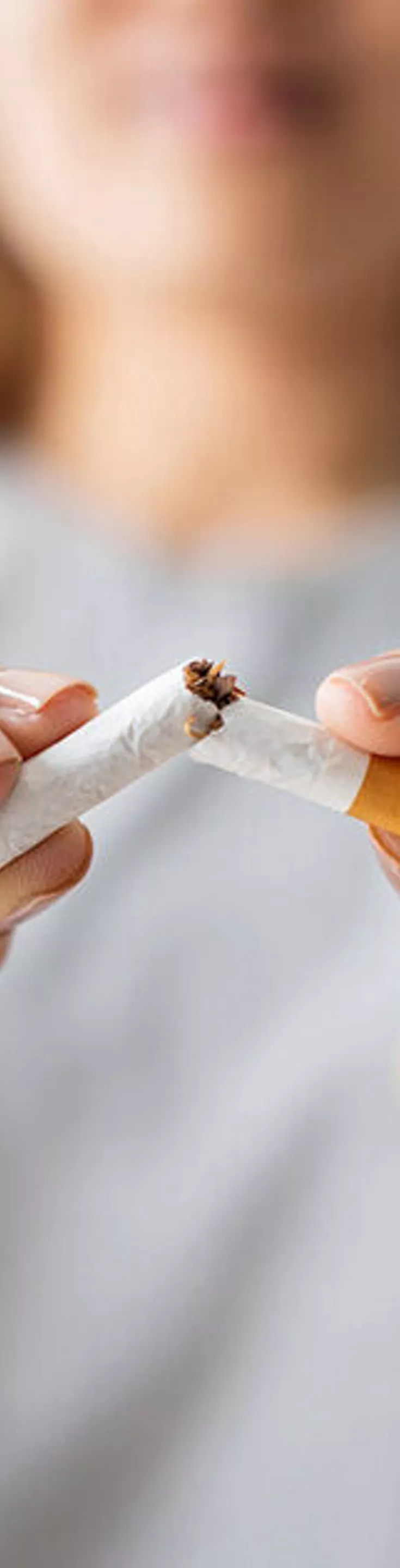 A woman is shown in close-up, breaking a cigarette in half with her hands. The focus is on the cigarette and her hands, symbolizing the act of quitting smoking. The background is softly blurred, emphasizing the subject and creating a neutral, supportive atmosphere. The woman appears to be an adult, and her expression is determined. No visible text or numbers are present in the image.