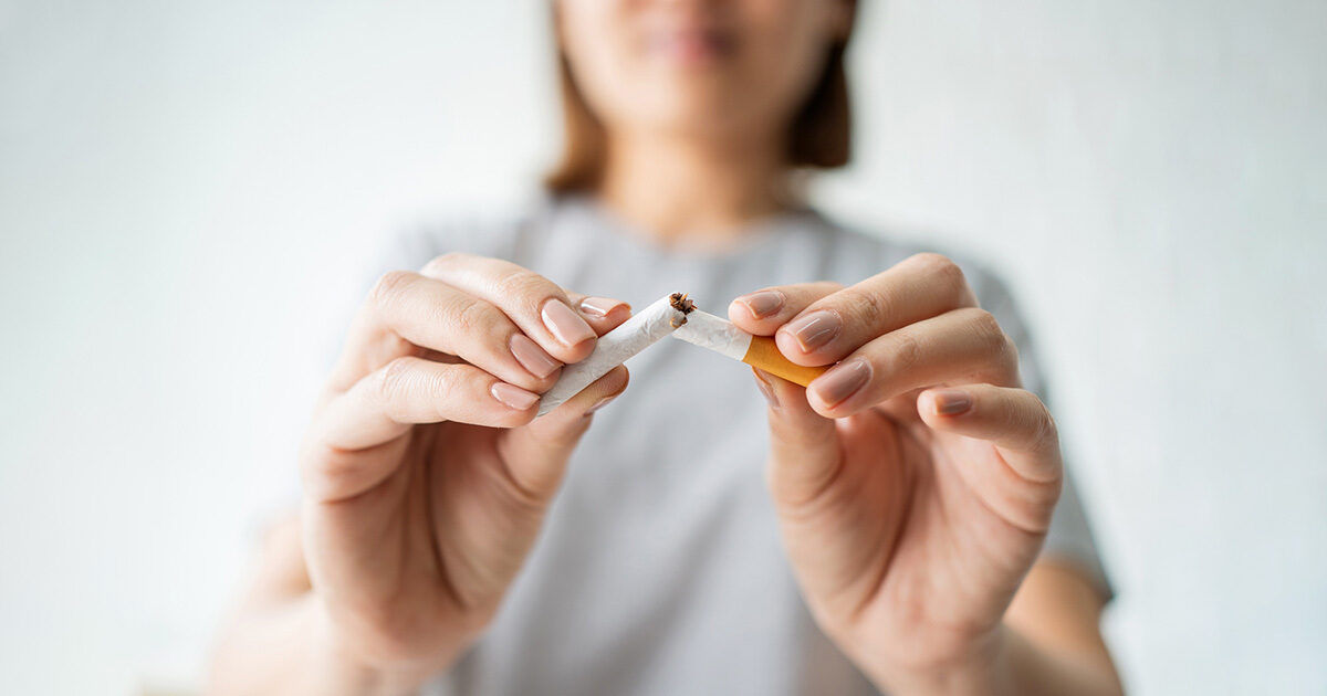 A woman is shown in close-up, breaking a cigarette in half with her hands. The focus is on the cigarette and her hands, symbolizing the act of quitting smoking. The background is softly blurred, emphasizing the subject and creating a neutral, supportive atmosphere. The woman appears to be an adult, and her expression is determined. No visible text or numbers are present in the image.