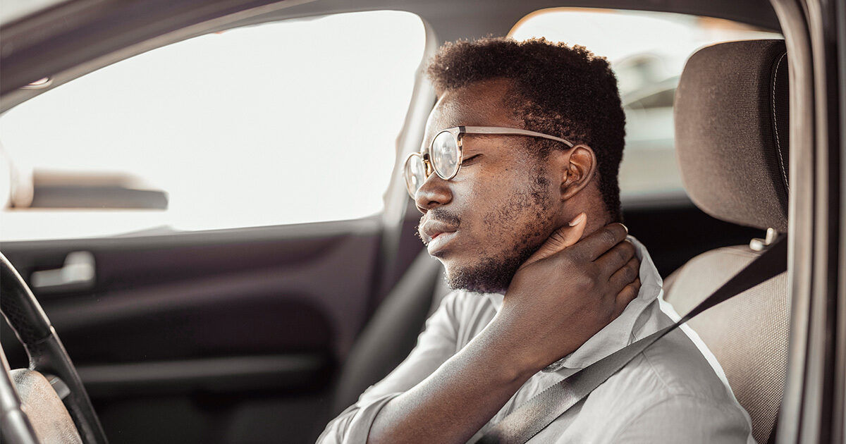A man sits in the driver's seat of a car, holding his neck with one hand as if experiencing pain or discomfort. The interior of the vehicle is visible, with the seatbelt fastened and sunlight streaming through the window. The scene suggests a possible injury or strain, commonly associated with driving or sudden movement. The overall mood is serious and reflective.