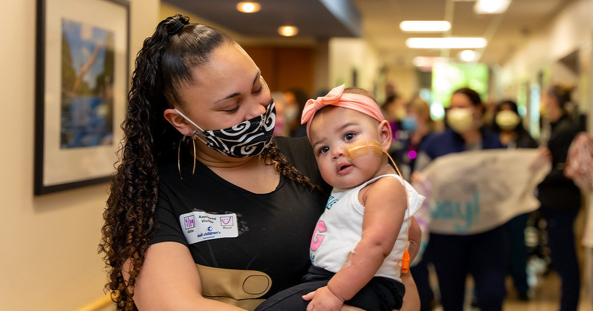 A woman with long curly hair holds a baby wearing a headband in a brightly lit hallway. The setting appears to be a public or community space, with several people and children visible in the background. The woman wears a black shirt and a name tag, and the baby is dressed in a white top. The mood is lively and social, with warm lighting and a casual atmosphere. Wall art and signage are visible, adding context to the indoor environment.