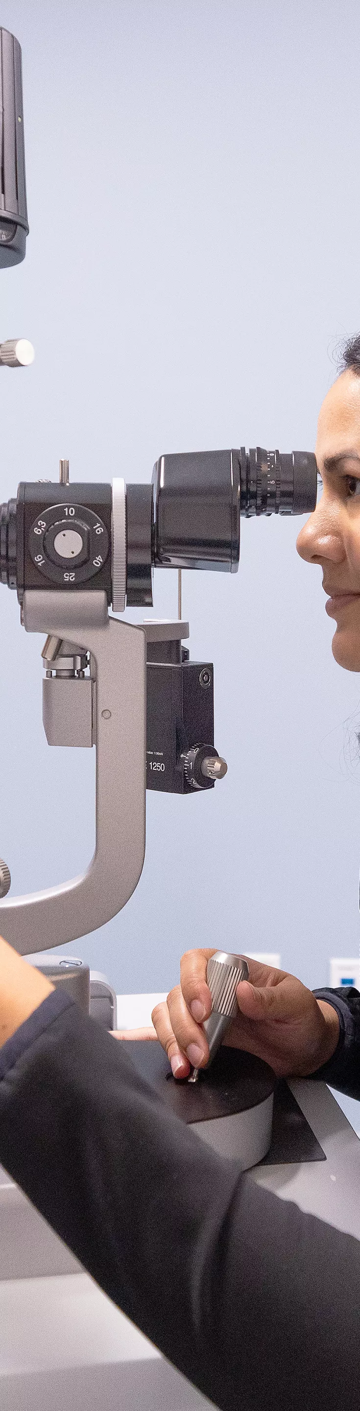 An adult male patient undergoes an eye examination using a slit lamp microscope in a clinical setting. A female ophthalmologist operates the equipment, focusing on the patient's eye health. 