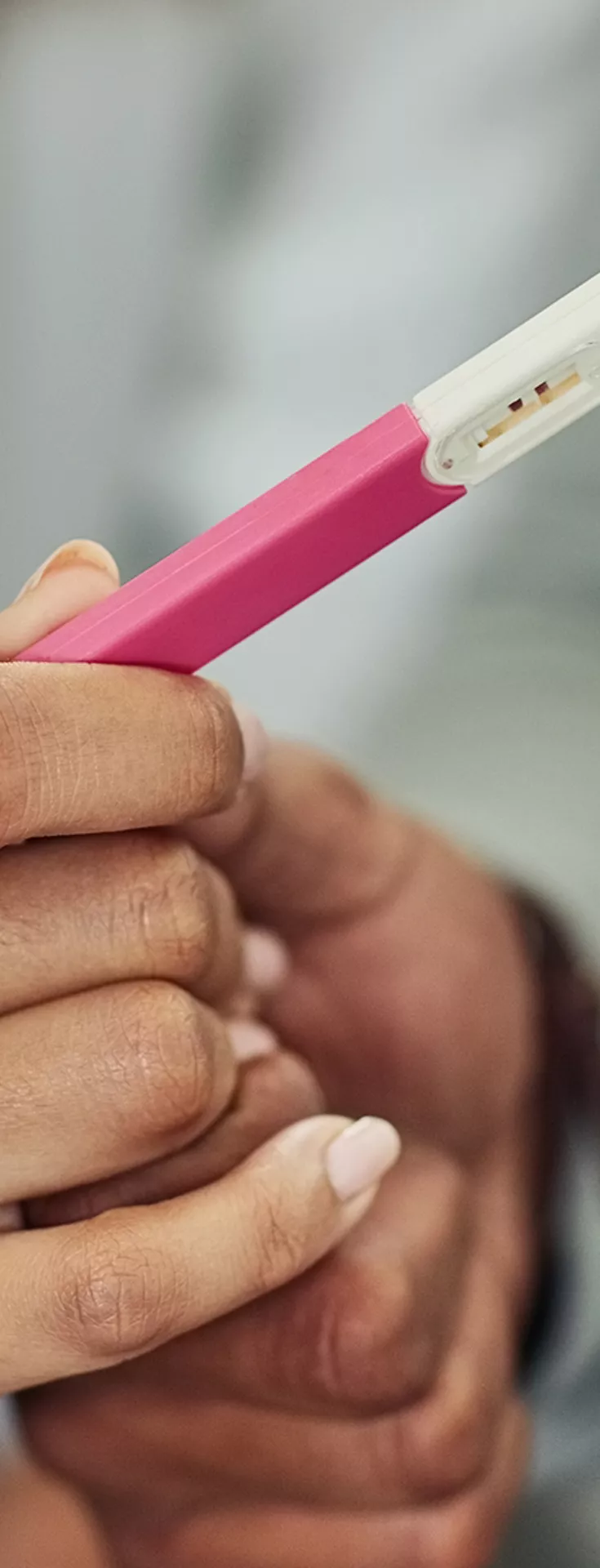 The image shows two people holding a pink and white pregnancy test stick together. The setting appears to be indoors, with the focus on their hands and the test device. The mood is intimate and anticipatory, with soft lighting and neutral tones. 
