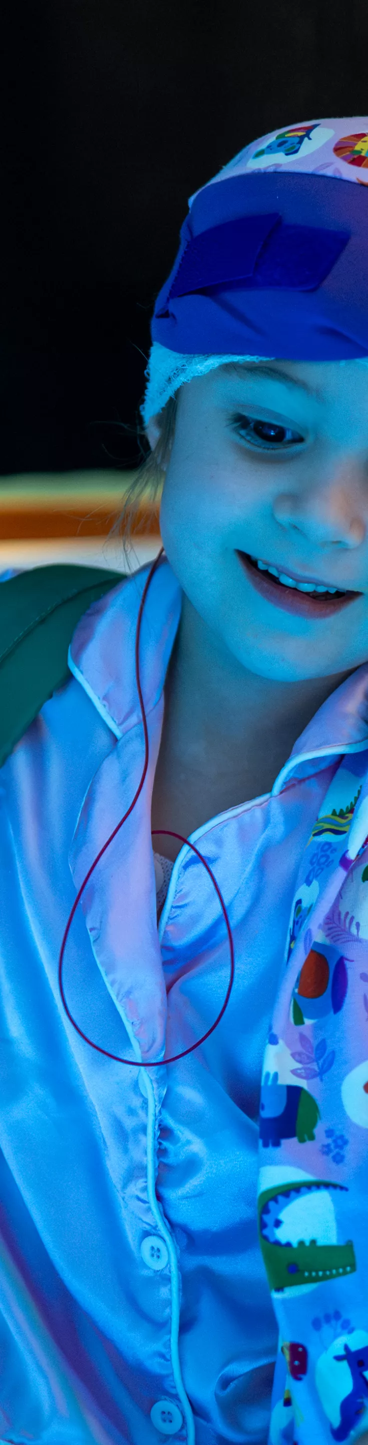 A young child sits in a sensory room, surrounded by glowing blue fiber optic strands. The child is wearing a colorful headscarf and light pink clothing, holding one of the illuminated strands. The environment is calming and therapeutic, with soft lighting and a relaxed atmosphere. A hospital wristband is visible on the child's arm.