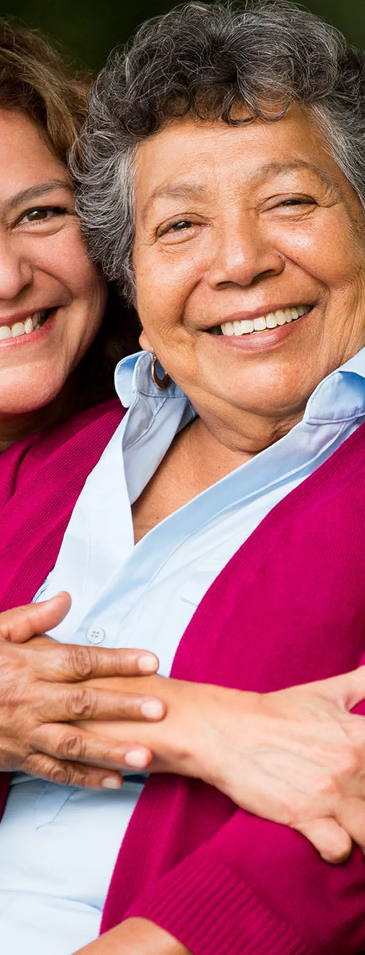 Two women are seated closely together on a wooden park bench, sharing a warm embrace. The setting is an outdoor park with lush greenery and trees in the background, creating a peaceful and natural atmosphere. One woman wears a red cardigan while the other is in a light-colored top, highlighting a moment of affection and support. The overall mood is caring and intimate, with soft, natural lighting.