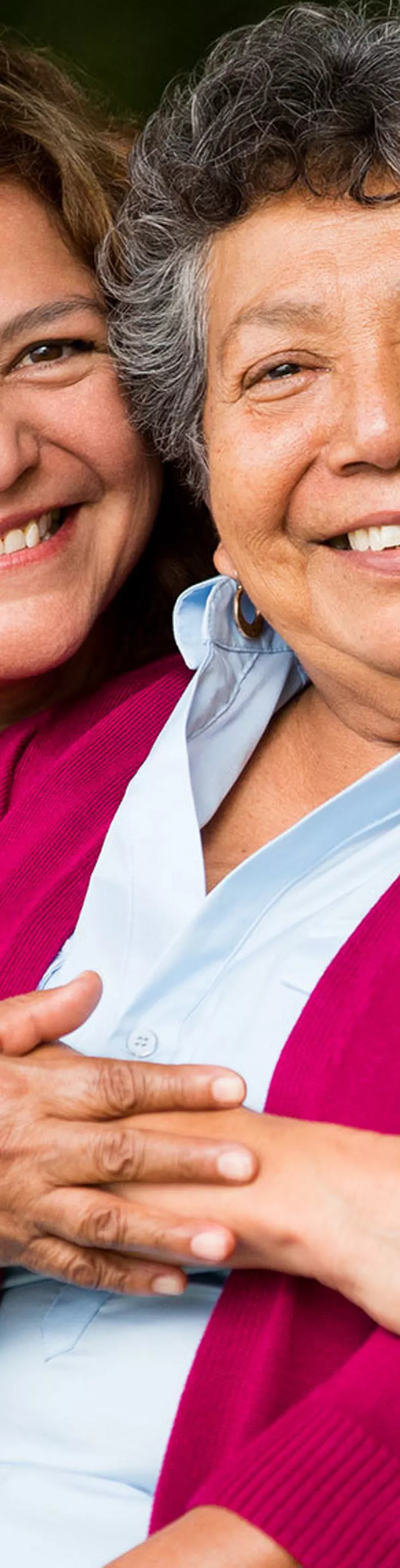 Two women are seated closely together on a wooden park bench, sharing a warm embrace. The setting is an outdoor park with lush greenery and trees in the background, creating a peaceful and natural atmosphere. One woman wears a red cardigan while the other is in a light-colored top, highlighting a moment of affection and support. The overall mood is caring and intimate, with soft, natural lighting.