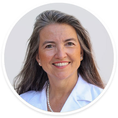 A woman with long brown and gray hair is pictured wearing a white coat and a pearl necklace. The background is a neutral light gray, creating a clean and professional look. The subject appears to be in a formal or business setting, possibly related to healthcare or academia. No visible text or numbers are present in the image.