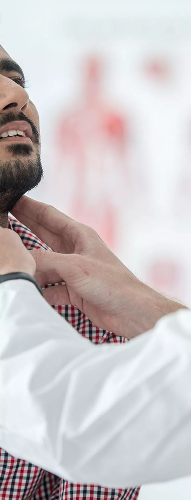 A healthcare professional in a white coat is examining the throat of an adult male patient wearing a red and white checkered shirt. The setting appears to be a bright, modern medical office with anatomical charts in the background. 