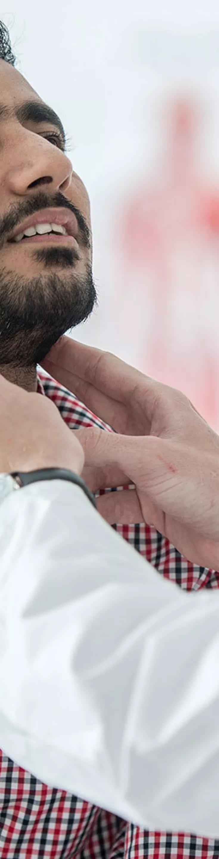 A healthcare professional in a white coat is examining the throat of an adult male patient wearing a red and white checkered shirt. The setting appears to be a bright, modern medical office with anatomical charts in the background. 