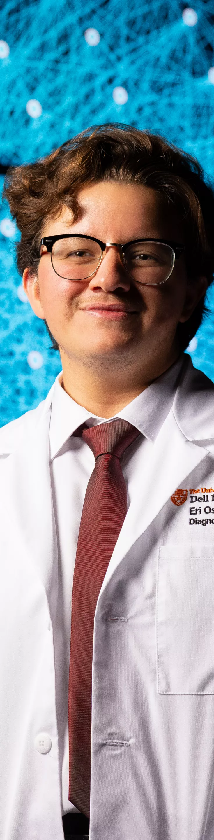 A person in a white medical coat stands in front of a large digital screen displaying a complex network visualization with blue and red lines. The individual appears to be a young adult male, dressed formally with a tie, suggesting a professional or academic setting. The background features a high-tech, scientific atmosphere, emphasizing data analysis or medical technology. Some text is visible on the coat, indicating credentials or department information.