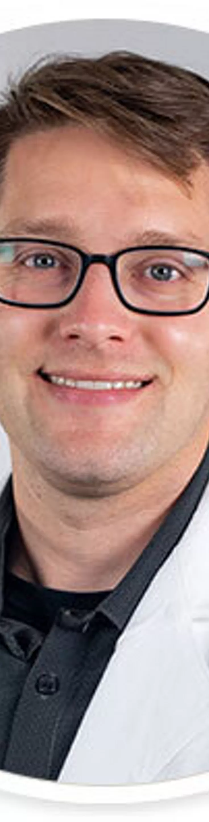 A man is pictured wearing a white lab coat over a black collared shirt, suggesting a professional or medical setting. The background is plain and neutral, focusing attention on the subject's attire. The image is presented in a circular frame, giving it a clean and modern look. No visible text or numbers are present.