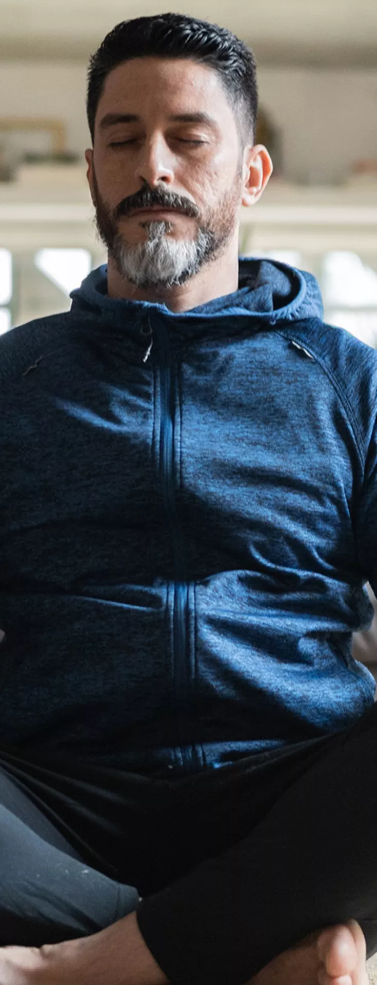 A person is seated cross-legged on a carpeted floor in a modern living room. He is wearing a dark blue hoodie and black pants, with a neutral and relaxed posture. The background features a grey sofa with a plaid blanket, large windows, and soft natural lighting. 