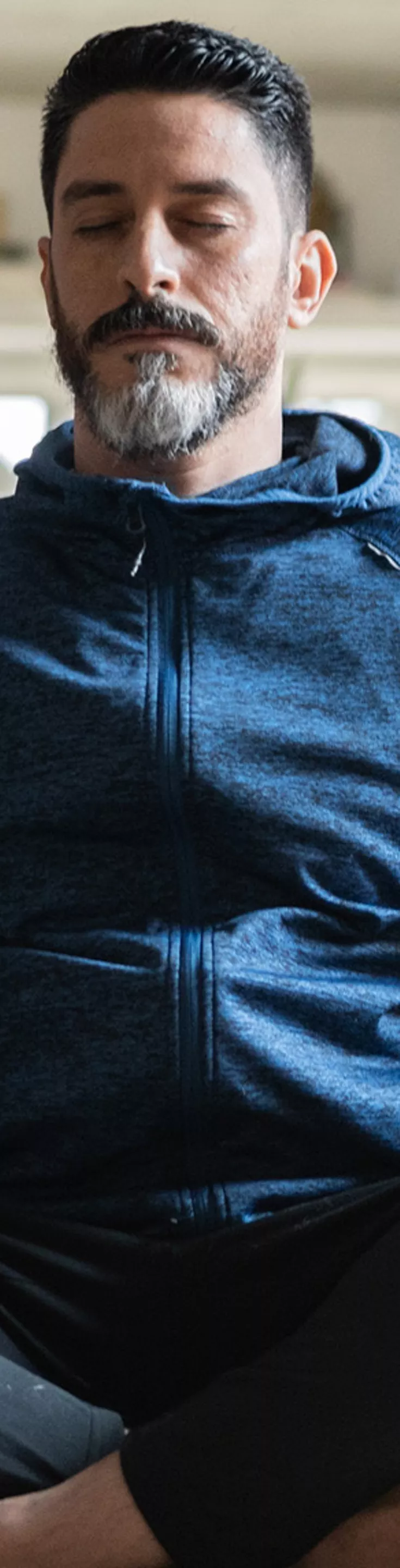 A person is seated cross-legged on a carpeted floor in a modern living room. He is wearing a dark blue hoodie and black pants, with a neutral and relaxed posture. The background features a grey sofa with a plaid blanket, large windows, and soft natural lighting. 