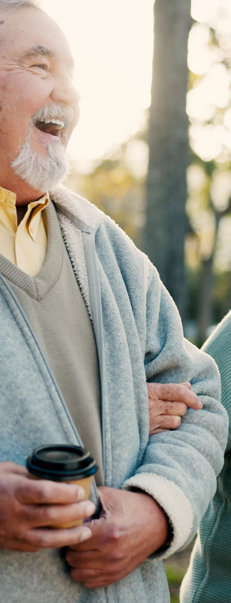 An elderly couple is seen walking arm in arm in a park setting, each holding a takeaway coffee cup. The man wears a light gray zip-up jacket over a yellow shirt, while the woman is dressed in a teal sweater. The background features trees and soft sunlight, creating a warm and relaxed atmosphere. 