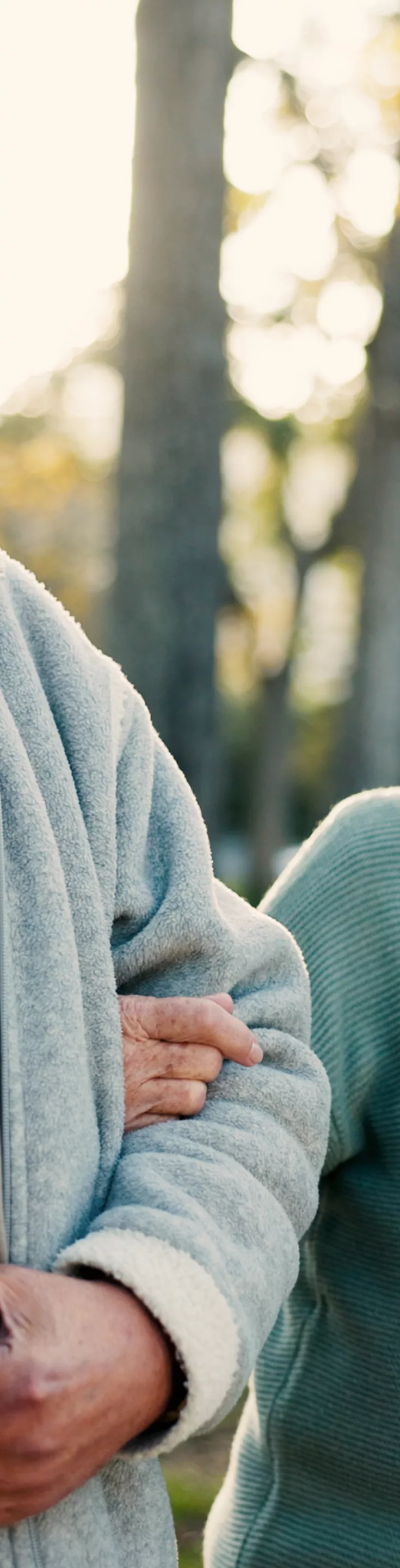 An elderly couple is seen walking arm in arm in a park setting, each holding a takeaway coffee cup. The man wears a light gray zip-up jacket over a yellow shirt, while the woman is dressed in a teal sweater. The background features trees and soft sunlight, creating a warm and relaxed atmosphere. 